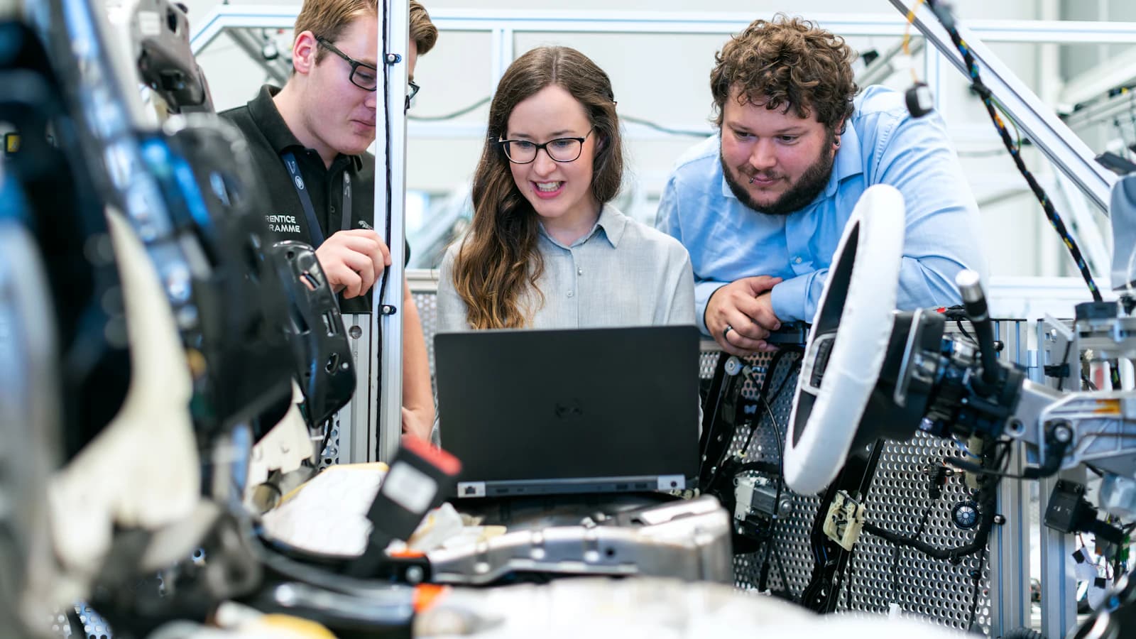 Three engineers, including an apprentice programme participant, gathered around a laptop while reviewing test data next to a wiring harness and robotics rig in a lab